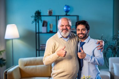 Hindistan 'ın kıdemli babası genç oğlunun kravat bağlamasına yardım ederken modern evdeki göreve hazırlanıyor, kameraya birlikte gülümsüyor, işten önce kendinden emin profesyonel bir adamı destekleyen deneyimli bir ebeveyn.