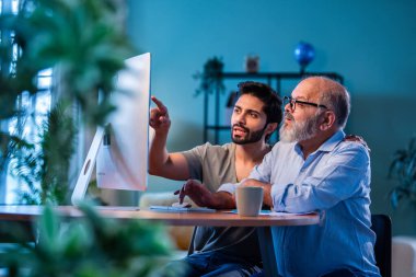 Hindistanlı genç oğul, babaya evde bilgisayar kullanmayı öğretiyor. Yaşlı baba, dijital teknolojiyi gülümseyerek öğrenirken, modern oturma odasında internete göz gezdiriyor.