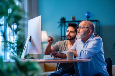 Hindistanlı genç oğul, babaya evde bilgisayar kullanmayı öğretiyor. Yaşlı baba, dijital teknolojiyi gülümseyerek öğrenirken, modern oturma odasında internete göz gezdiriyor.