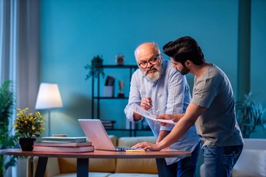 Hintli genç oğul ve kıdemli baba online vergi beyannamesini birlikte dolduruyorlar. Modern evdeki masada otururken mali raporları, yıllık gelir beyannamelerini tartışıyorlar.