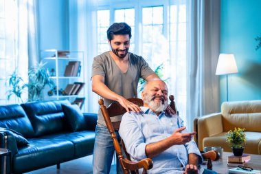 Hintli genç oğlu, modern oturma odasında sallanan sandalyeye yaslanıp yaşlı bir ebeveyn olarak ilgi, şefkat ve saygı göstererek yaşlı babaya masaj yapıyor. Evde rahatlatıcı bir omuz rahatlığı yaşıyor.