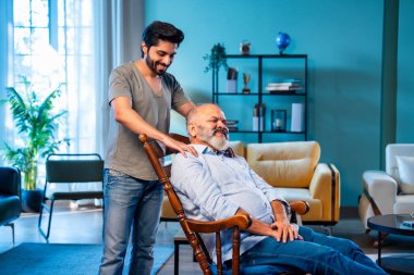 Hintli genç oğlu, modern oturma odasında sallanan sandalyeye yaslanıp yaşlı bir ebeveyn olarak ilgi, şefkat ve saygı göstererek yaşlı babaya masaj yapıyor. Evde rahatlatıcı bir omuz rahatlığı yaşıyor.