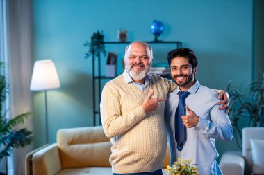 Hindistan 'ın kıdemli babası genç oğlunun kravat bağlamasına yardım ederken modern evdeki göreve hazırlanıyor, kameraya birlikte gülümsüyor, işten önce kendinden emin profesyonel bir adamı destekleyen deneyimli bir ebeveyn.