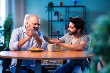 Hintli genç çocuk, modern oturma odasında yaşlı babasıyla ciddi bir konuşma yapıyor. Duygusal aile tartışması iki nesil arasında rehberlik, güven ve anlamlı bir bağ paylaşımı.