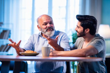 Hintli genç oğlu, modern oturma odasındaki babasıyla neşeyle konuşuyor. İki kuşak arasında iç açıcı bir aile sohbeti. Tavsiyelerini, bilgeliğini, mutluluğunu ve gülümsemesini paylaşıyor.