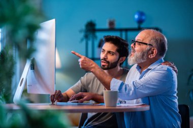 Hindistanlı genç oğul, babaya evde bilgisayar kullanmayı öğretiyor. Yaşlı baba, dijital teknolojiyi gülümseyerek öğrenirken, modern oturma odasında internete göz gezdiriyor.