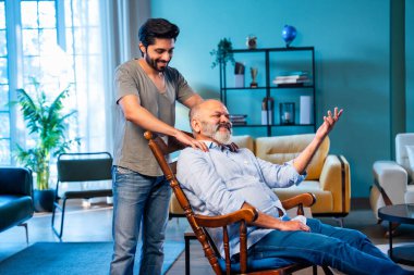 Hintli genç oğlu, modern oturma odasında sallanan sandalyeye yaslanıp yaşlı bir ebeveyn olarak ilgi, şefkat ve saygı göstererek yaşlı babaya masaj yapıyor. Evde rahatlatıcı bir omuz rahatlığı yaşıyor.