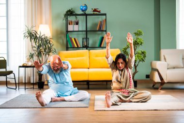 Hintli son sınıf çifti, modern oturma odasında paspasın üzerinde otururken evde Janushirasanaor yogası yapıyor.