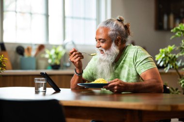 Hintli orta yaşlı adam evde yemek yerken akıllı telefon makaraları izliyor, şık sakallı erkek kahvaltı masasında poha yiyor ve modern mutfak yaşam tarzında internet yayını veya komedi videoları izliyor.