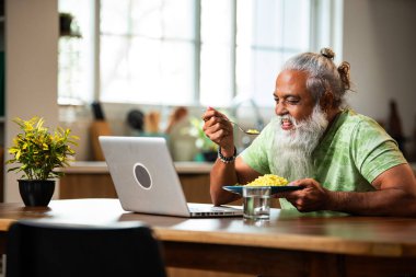 Hintli orta yaşlı adam evde yemek yerken dizüstü bilgisayar izliyor, şık sakallı erkek kahvaltı masasında poha yiyor ve modern mutfak iç dekorasyonunda internet dizisi veya komedi filmi seyrediyor.