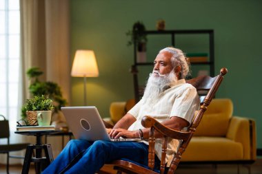 Hintli orta yaşlı sakallı adam dizüstü bilgisayarda çalışıyor evdeki rahat koltukta oturuyor, modern iç mekanda kablosuz dijital teknoloji yaşam tarzıyla modern bir şekilde bilgisayarda yazan olgun bir erkek.