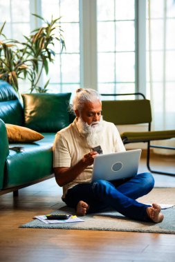 Hindistan 'ın orta yaşlı sakallı adamı modern evdeki halıda oturan nakit Hint rupilerini sayıyor. Evin içinde kağıt para birimiyle ev finansmanı ve bütçesini yöneten olgun bir erkek.