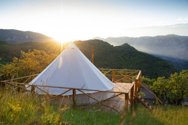 beautiful panorama of glamping house tent in the morning sun light on wooden deck with fence and foggy mountain range scenery in the background clipart
