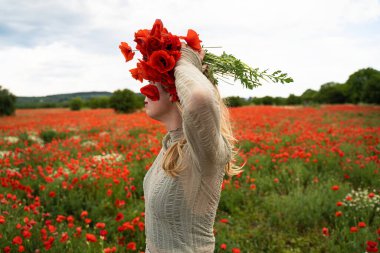 Kafasında kırmızı çiçeklerle dolu güzel bir tarlada elinde bir buket haşhaş tutan açık renkli elbiseli sarışın beyaz kadının yakın plan portresi.
