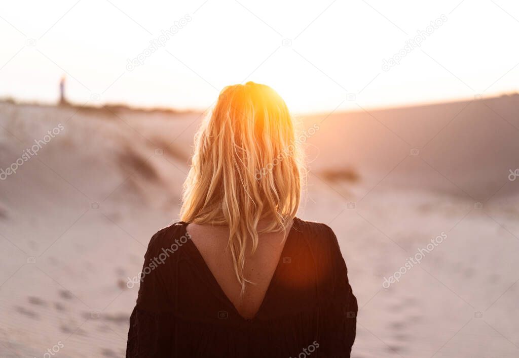 rear view sunset portrait of Caucasian blonde young beautiful woman in black dress and sun shining in hair standing with back at the camera in front of sand dune