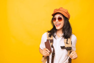 Asyalı kadın sırt çantalı, yazın seyahat promosyonuna bilet almış, genç kız gülümsüyor, elinde bavul ve pasaport, stüdyo sarısı arka planda..