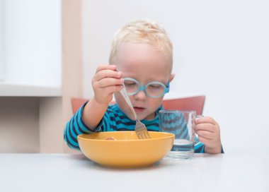 Gözlüklü erkek bebek önünde sarı bir tabak bir masada oturur