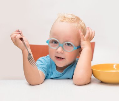 Gözlüklü erkek bebek önünde sarı bir tabak bir masada oturur
