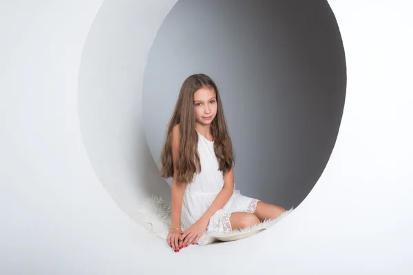 Uzun dalgalı kahverengi saçlı kızı beyaz bir duvar kapı aralığında oturur