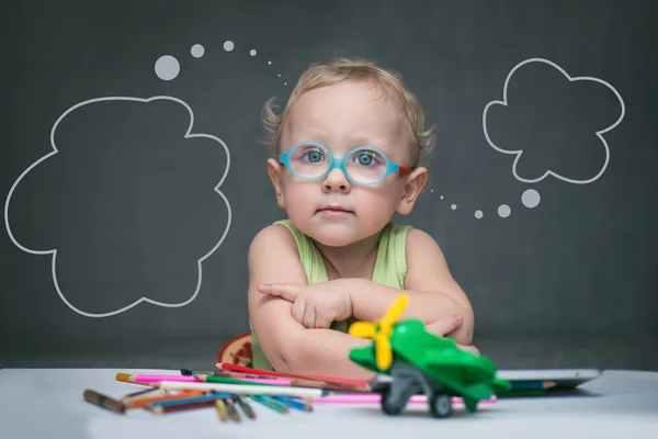 Маленький мальчик в очках сидит за столом, на котором разбросаны мелки и игрушки — стоковое фото