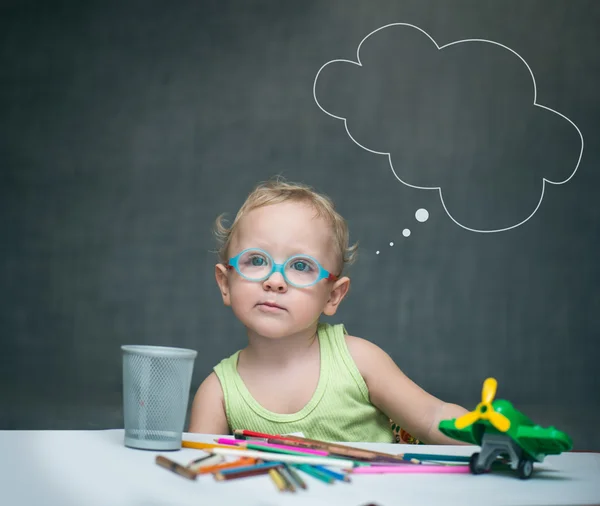 Gözlüklü küçük çocuk üzerinde boya kalemi ve oyuncaklar dağılmış bir masada oturur