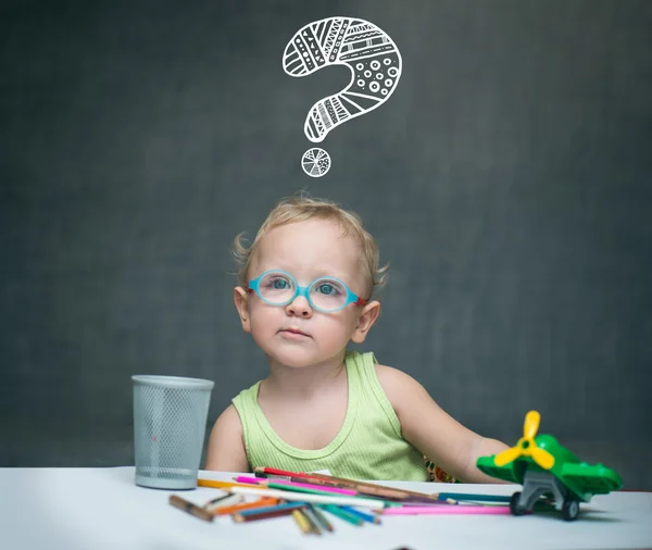 Gözlüklü küçük çocuk üzerinde boya kalemi ve oyuncaklar onun kafa asmak üzerinde dağılmış bir masada oturur