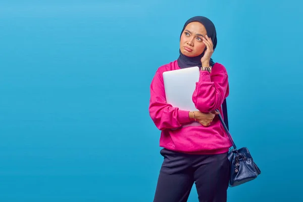 Photo of sad asian woman wearing pink jacket carrying laptop looking at empty space with hands holding head isolated on blue background