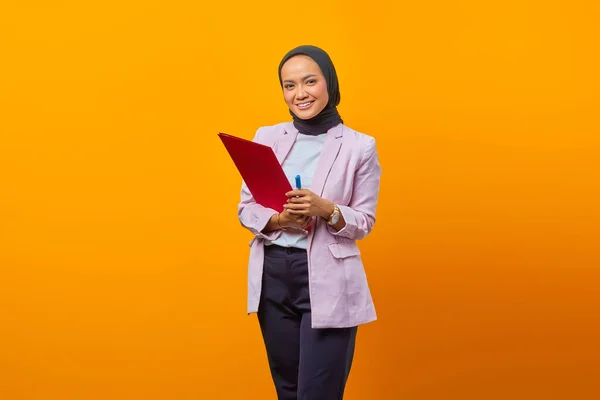 Beautiful asian woman holding folders and pen while looking at camera with smiling face on yellow background