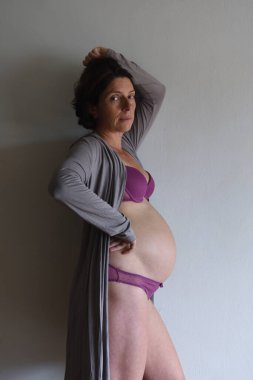 Duvara yaslanmış kameraya bakan hamile bir kadın.