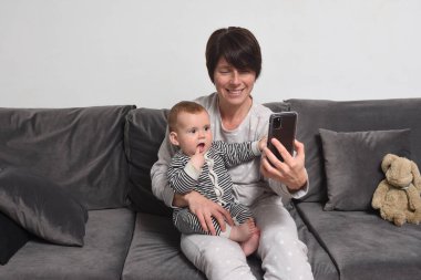 Anne ve oğlu kanepede oturuyor, anne bebeğe akıllı telefonu gösteriyor.