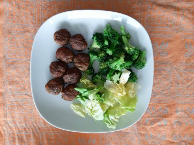 Kaynamış brokoli ile yeşil salata yaprakları ve beyaz tabakta servis edilen köfteler.