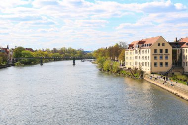 Regenburg, Almanya, 18 Nisan 2022: İlkbaharda Tuna Nehri kıyısındaki manzaralı caddede yürüyen insanlarla birlikte şehir manzarası
