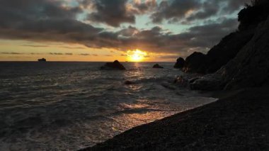 Bogliasco İtalya 'sındaki kara bulutların altında kumlu kayaları yıkayan sakin okyanus dalgaları üzerinde dramatik bir gün batımı..