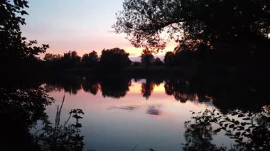 Altın güneş, Regensburg yakınlarındaki sakin göl suyuna yansıyan orman siluetlerinin ardında batıyor.