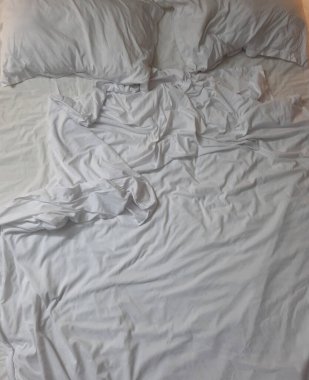 Yumuşak beyaz yatak örtüleri buruşuk çarşaflar ve yumuşak yastıklar, rahat ve cezbedici bir atmosfer yaratıyor iyi aydınlatılmış bir yatak odasında, huzurlu bir ortamda rahatlamak ve rahatlamak için mükemmel.