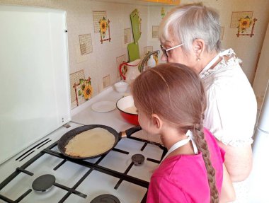 Bir büyükanne ve torun mutfakta krep yapıyorlar, tava ve gaz sobasıyla. Pembe tişörtlü ve beyaz önlüklü bir kız süreci izliyor..