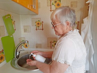 Yaşlı bir kadın mutfak lavabosundaki musluktan akan suyun altında ellerini sabunla yıkıyor..