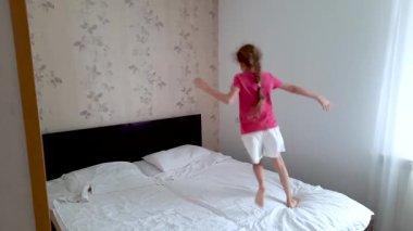 Beyaz çarşaflı bir yatakta mutlu bir şekilde koşan bir kız. Pencereli bir ev ya da otel odası. Aile kavramları, oyun ve seyahat.