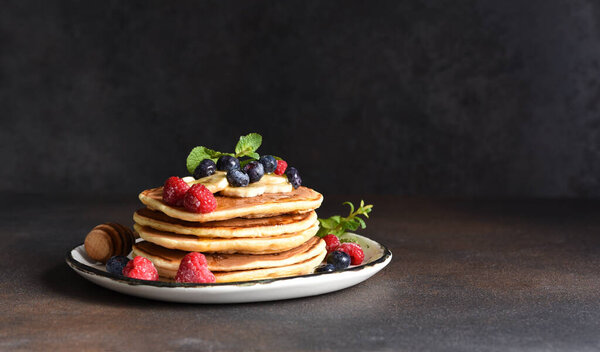 Homemade pancakes with banana, raspberries, blueberries and honey on a dark concrete background.