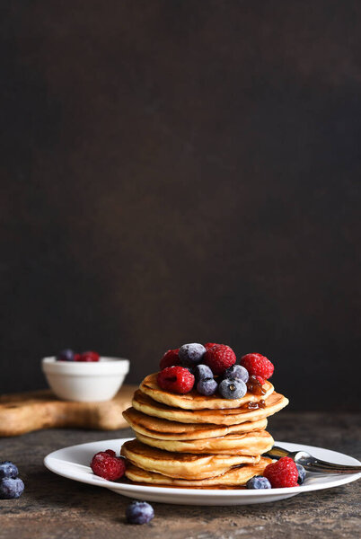 Delicious homemade pancakes in a plate with berries: raspberries, blueberries and maple syrup on a concrete background. With copy space.