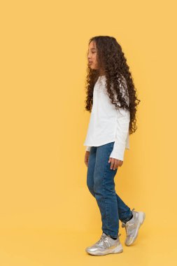Sarı arka planda beyaz gömlek ve kot pantolon giyen, yürüyen, koyu tenli, kıvırcık saçlı Afrikalı bir kızın stüdyo fotoğrafı..
