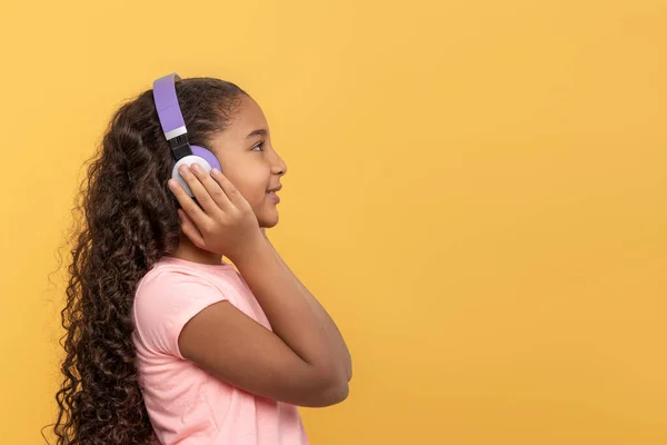 近景肖像一个年轻的深色皮肤女孩 长发卷曲 身穿粉色衬衫 带着黄色背景的紫色耳机听音乐 — 图库照片