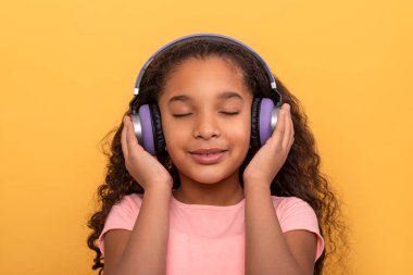  Uzun kıvırcık saçlı, pembe tişörtlü, mor kulaklıklı, gözleri sarı bir arka planda kapalı müzik dinleyen genç bir kızın yakın portresi.