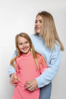 Mavi ve pembe süveter giyen mutlu anne ve kızın yarım boy portresi stüdyoda beyaz bir arka planda birbirlerine sarılıyorlar.
