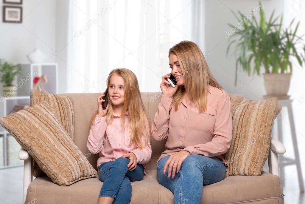 Shot of a mom and daughter of blondes with long hair wearing pink shirts and jeans sitting on the couch at home and having fun talking on the phone. Daughter imitates mom's behavior