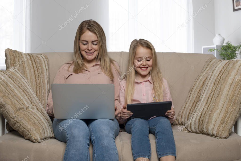 A shot of a mom and daughter of blondes with long hair wearing pink shirts and jeans sitting on the couch at home with a laptop and a tablet.  Daughter imitates mom's behavior