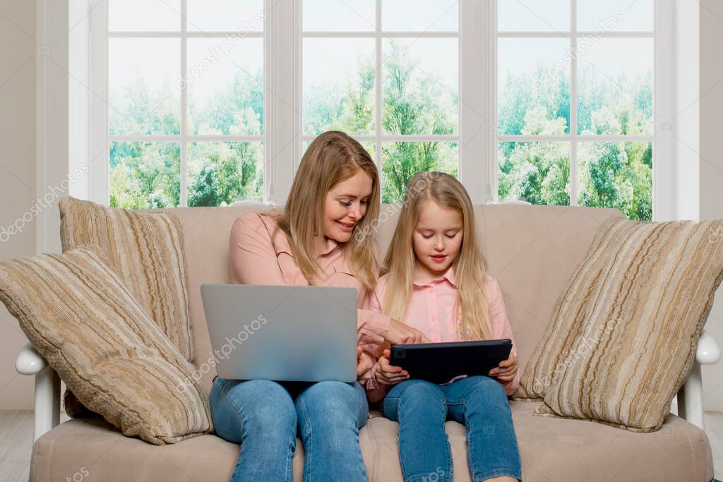 A shot of a mom and daughter of blondes with long hair wearing pink shirts and jeans sitting on the couch at home with a laptop and a tablet.  Daughter imitates mom's behavior