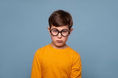 Yuvarlak gözlüklü, sarı tişörtlü, mavi arka planda bir şeyden memnun olmayan komik bir çocuğun stüdyo fotoğrafı.