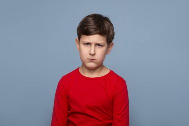 Kırmızı tişörtlü genç bir çocuğun, mavi arka plana karşı tatminsiz bir görüntüsü.