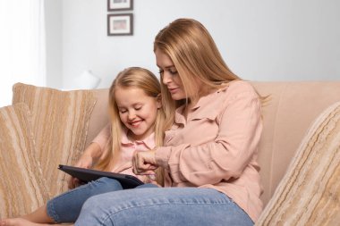 Uzun saçlı, pembe gömlekli ve kot pantolonlu bir anne ve kızının tabletle evdeki kanepede oturduğu bir fotoğraf. Evde birlikte eğleniyorlar..